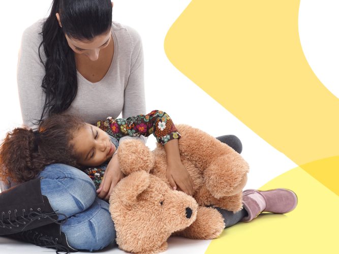 A child holds a teddy bear and is hugged by a parent